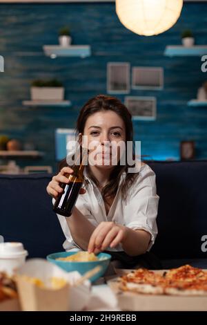 Porträt einer Frau, die die Kamera mit der Bierflasche in der Hand hält und Kartoffelchips von der Schüssel am Tisch mit großer Pizza und Abendessen zum Mitnehmen isst. Büroangestellter auf der Couch, der sich nach dem Essen zum Mitnehmen entspannt. Stockfoto