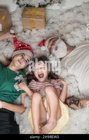 Weihnachtsfoto der glücklichen Familie. Neujahrsporträt von Vater, Mutter und Tochter in weihnachtlichen roten Hüten, lächelnd und im weißen Kunstschnee liegend. Stockfoto