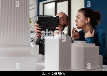 Projektingenieure arbeiten zusammen und betrachten ein Tablet mit Bauplänen, die im modernen Architekturbüro am Schreibtisch sitzen. Zwei Architekten machen Teamarbeit neben dem Tisch mit Gebäude maquette. Stockfoto