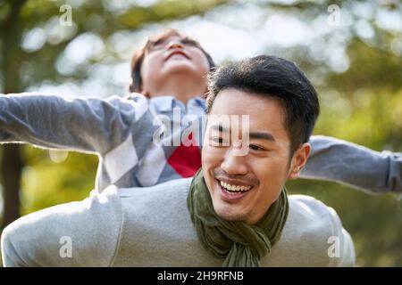 asiatischer Vater trägt Sohn auf dem Rücken mit Spaß im Freien im Park Stockfoto