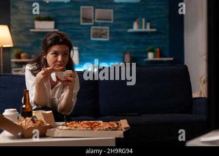 Frau, die sich auf der blauen Couch lässig kleidete, aß asiatische Nudeln zum Mitnehmen mit Essstäbchen und schaute sich die Abendnachrichten im Fernsehen an. Person mit entspannendem Take-out chinesischen Fast Food nach Hause Lieferservice Menü. Stockfoto