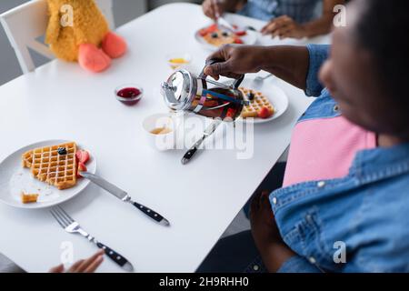 Blick aus der Vogelsicht auf die verschwommene afroamerikanische Frau, die beim Frühstück mit der Familie Tee in die Nähe von leckeren Waffeln mit Beeren goss Stockfoto