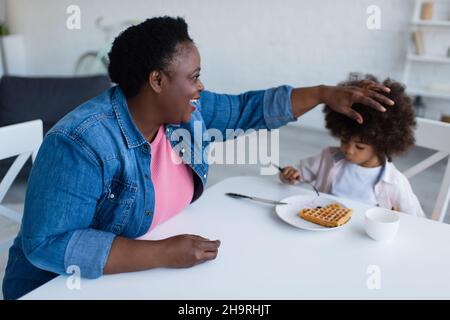 Glückliche afroamerikanische Oma, die lockiges Haar der Enkelin berührte, die zum Frühstück Waffel aß Stockfoto
