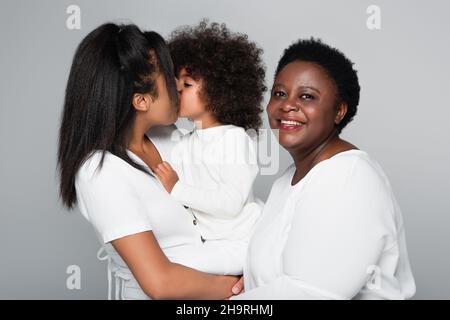Junge afroamerikanische Frau, die ihre Tochter im mittleren Alter küsst, lächelt mit der Kamera, die auf Grau isoliert ist Stockfoto