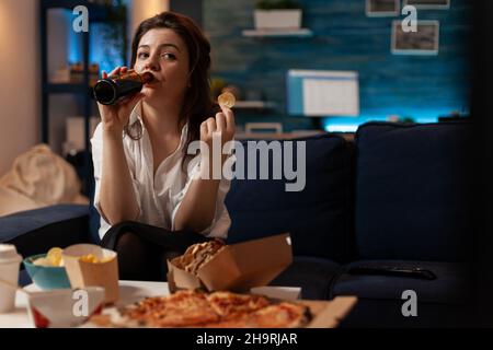 Frau bekleidet Casual trinkt Bier aus der Flasche, während sie einen Kartoffelchip in der Hand hält und sich die TV-Comedy-Sitcom-Show anschaut. Person, die auf der Couch sitzt und vor dem Tisch vor dem fernseher mit Fast Food schaut. Stockfoto