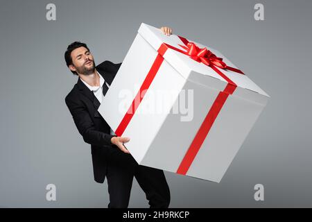 Bärtiger Mann in Anzug mit riesiger Geschenkbox isoliert auf grau Stockfoto