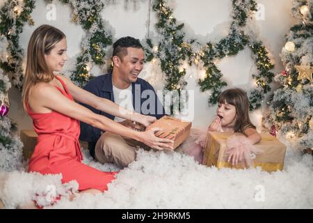 Neujahrsfoto von glücklicher gemischter Familie mit niedlicher Tochter. Eltern schenken ihrer Tochter Weihnachtsgeschenke. Mädchen überrascht von Geschenk Geschenkbox. Stockfoto