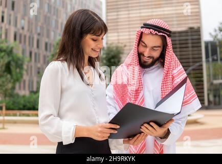 Glücklicher arabischer Mann in keffiyeh lächelt und liest Papiere in Ordner während eines Geschäftstreffens mit jungen Managerin in der Stadt Stockfoto