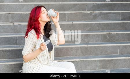 Junge Frau, die auf der Treppe im Freien sitzt und aus einem zusammenklappbaren Becher aus Silikon trinkt, wiederverwendbarer Coffee-to-go-Becher. Stockfoto