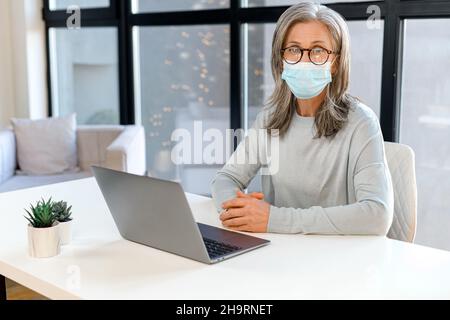 Seious reife grauhaarige Geschäftsfrau trägt Maske im modernen Büro schaut auf die Kamera, tippt auf den Laptop, schreibt eine E-Mail. Schutzmaßnahmen für Büromaterial während einer Pandemie Stockfoto