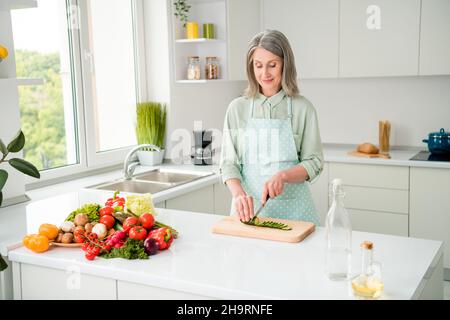 Porträt einer attraktiven, fröhlich fokussierten grauhaarigen Frau, die hausgemachte Abendessen Bio-Farm-Produkte zu Hause zubereitet, hellweiß im Innenbereich Stockfoto