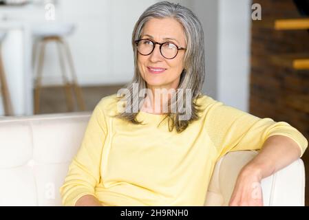 Nahaufnahme des Porträts einer modernen älteren Frau mit stilvoller Brille blickt auf die Kamera und lächelt, eine ältere Frau, die sich zu Hause ausruht und ein ruhiges Wochenende verbringt Stockfoto