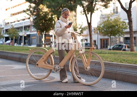 Stilvolle Frau mit Holz Öko-Fahrrad steht auf dem Bürgersteig und Surfen Handy im Park der modernen Stadt. Stockfoto