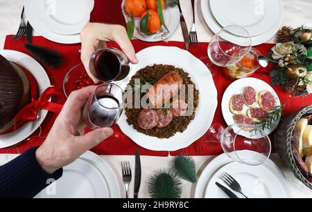 Italienisches Weihnachtsessen. Flach auflegte Hände, die mit Gläsern Rotwein toasten. Cotechino mit Linsen, Vorspeise, Obst und Panettone.Familientreffen. Stockfoto