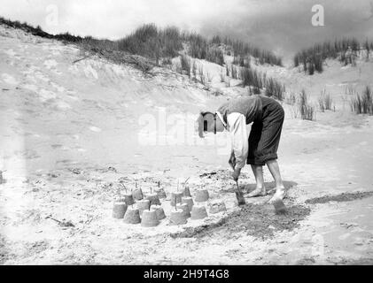 1950s, historisch, draußen an einem Sandstrand, an einigen Sanddünen, ein Mann mit seiner Nadelstreifenhose bis zu den Knien gerollt und einem ärmellosen Pullover über seinem Hemd, der mit einem kleinen Kinderspaten endet, Sandburgen baut, alle mit kleinen Stöcken auf ihnen platziert, England, Großbritannien. Stockfoto