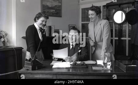 1950s, ein historischer Geschäftsmann, der an seinem Schreibtisch in seinem Büro saß, neben dem eine Sekretärin am Telefon stand und eine gut gekleidete Assistentin, mit einem Füllfederhalter in der Hand, damit er einige Papiere unterschreiben konnte, England, Großbritannien. Stockfoto
