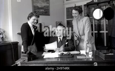 1950s, ein historischer Geschäftsmann, der an seinem Schreibtisch in seinem Büro saß, neben dem eine Sekretärin am Telefon stand und eine gut gekleidete Assistentin, mit einem Füllfederhalter in der Hand, damit er einige Papiere unterschreiben konnte, England, Großbritannien. Stockfoto