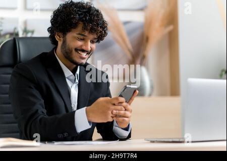 Online-Chat mit dem Smartphone. Zufriedener Inder, Top-Manager, Geschäftsinhaber sitzt an einem Schreibtisch, nutzt ein Mobiltelefon während der Arbeit, im Internet surfen, E-Mails beantworten, mit Freunden schreiben, lächelt Stockfoto