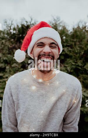 Mixed-Race-Mann lacht im Weihnachtsmütze und Pullover, die im Winter vor immergrünen Bäumen Weihnachtslichter um den Hals tragen Stockfoto