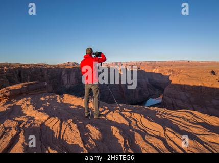 Glen Canyon National Recreation Area, Page, Arizona, USA. Fotograf am Cliff Edge beim Fotografieren des Colorado River im Horseshoe Bend, Sonnenaufgang. Stockfoto