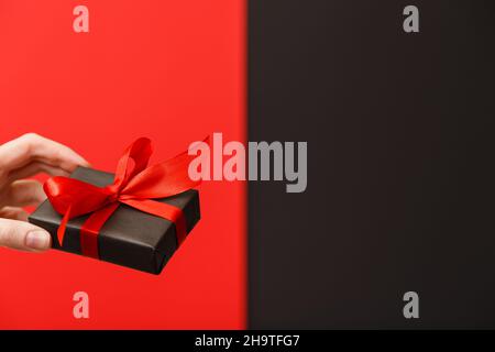 Weibliche Hand hält schwarze Geschenkbox mit roter Schleife über leuchtend reichen roten und schwarzen Hintergrund. Black friday Sale, Weihnachtsverkauf, Rabattkonzept. Valentinstag. Vatertag Stockfoto