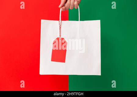 Weibliche Hand hält weiße leere Papier-Einkaufstasche mit Preisschild isoliert auf rotem und grünem Hintergrund. Black friday Verkauf, Weihnachtsverkauf, Rabatt, Shopping und Ökologie Konzept Stockfoto