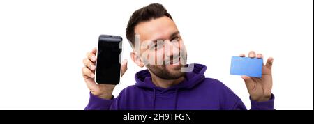 Ein charmanter Mann in einem violetten Hoodie demonstriert einen Telefonbildschirm und eine Bankkarte oder Visitenkarte oder eine Rabattkarte Stockfoto
