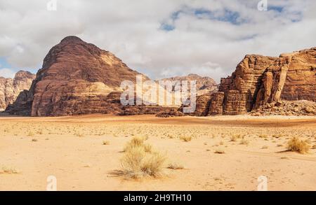 Felsige Massive auf roter Sandwüste, wenig trockener Grashaufen, wolkiger Himmel im Hintergrund, typische Landschaft im Wadi Rum, Jordanien Stockfoto