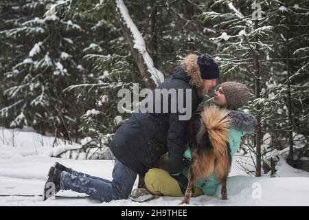 Verliebte Paare, die im Winterwald im Schnee Spaß mit dem Hund haben Stockfoto