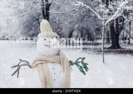 Glücklicher Schneemann im Freien im verschneiten Winterpark in gestricktem weißen Hut, Schal mit Tannenzweigen in den Händen gemacht. Saisonale Aktivitäten Stockfoto