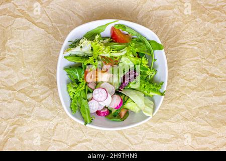 Draufsicht auf leuchtend frische grüne Salatblätter mit roten Tomaten, Gurken und Rettich in der weißen Schüssel auf hellem Hintergrund. Stockfoto