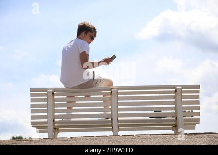 '20.06.2021, Deutschland, Niedersachsen, Hannover - der junge Mann sitzt auf der Rückseite einer Parkbank und schaut auf sein Handy. 00S210620D467CAROEX.JPG [MODELL Stockfoto