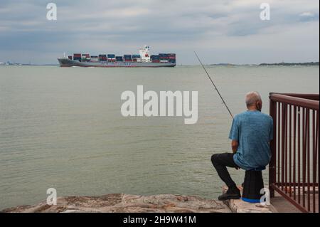 '09.10.2021, Singapore, , Singapur - Ein Mann sitzt am Ufer des Changi Beach Park und fischt, während ein Frachtschiff im Hintergrund vorbeifährt. 0SL211009D016 Stockfoto