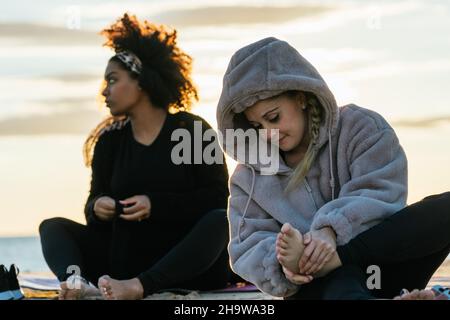 Zwei Frauen in warmen Kleidern sitzen bei Sonnenuntergang am Strand nach einem Yoga-Kurs Stockfoto