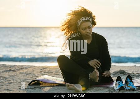 Eine Schwangerin sitzt am Strand und wischt sich während des Sonnenuntergangs den Sand von den Füßen Stockfoto