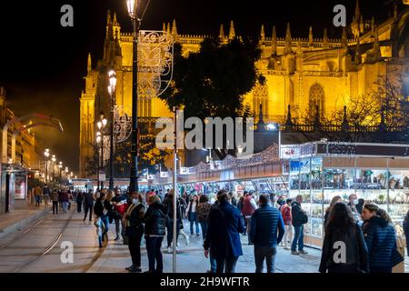 Nacht auf dem Weihnachtsmarkt, während Touristen und einheimische Spanier die Dekorationen mit der Kathedrale von Sevilla im Blick hinter sich genießen. Stockfoto