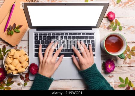 Frau mit Tasse Tee, Notizbuch, Essen, Blättern und Blumen mit Laptop auf hellem Holzboden Hintergrund Stockfoto