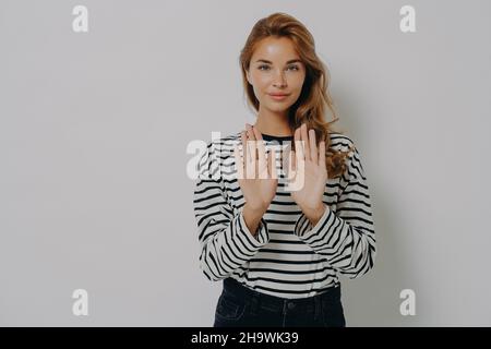 Junge selbstbewusste Frau, die ein Stoppschild gestikuliert und Nein sagt, während sie isoliert gegen eine graue Wand steht Stockfoto