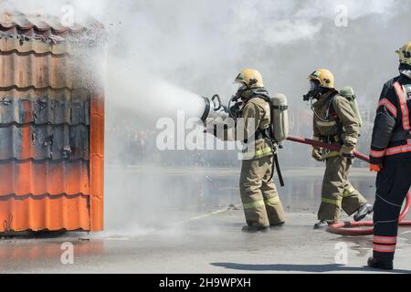 Gruppe von Feuerwehrleuten, die das Feuer aus dem Löschschlauch löschen, mit Löschwasser-Schaumstofffass mit Schaum während des Berufsurlaubs Firefighters Day Stockfoto