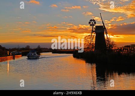 Ein Kreuzer auf dem Fluss Ant auf den Norfolk Broads vorbei an der Turf Fen Drainage Mill mit Herbstuntergang in der Nähe von Ludham, Norfolk, England, Großbritannien. Stockfoto