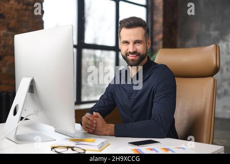 Optimistischer, männlicher Büroangestellter, der am Schreibtisch vor dem PC sitzt, die Kamera anschaut und lächelt, erfolgreicher junger Geschäftsmann im modernen Loft-Büro, Manager-ceo in eleganter Casual-Atmosphäre am Arbeitsplatz Stockfoto