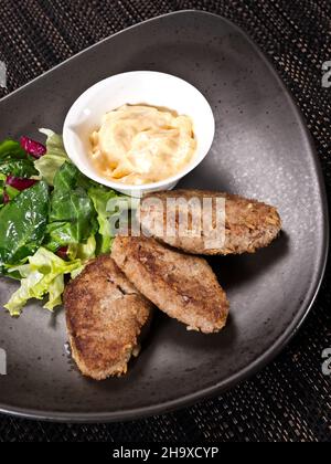 Gebratene Fleischkoteletts serviert mit cremiger Sauce und frischem grünen Salat auf einem dunklen Teller Stockfoto