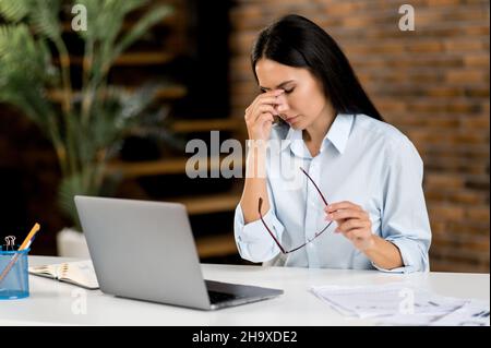 Kopfschmerzen und Migräne. Erschöpfte kaukasische junge Erwachsene Frau, Top-Manager, Büroangestellte, müde von der Arbeit, in Stress, Machte eine Pause von der Arbeit, braucht eine Pause, massiert den Nasenrücken, schließt die Augen Stockfoto