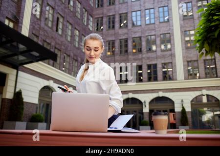 Erfolgreiche Geschäftsfrau mit Smartphone, das an einem Laptop im Freien arbeitet. Stockfoto