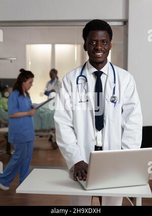 Porträt eines lächelnden afroamerikanischen Spezialisten-Therapeuten-Arztes, der bei der Gesundheitsbehandlung auf der Krankenhausstation Erfahrungen mit Krankheiten am Computer eingibt. Medizinisches Team, das den Patienten nach der Operation unterstützt Stockfoto