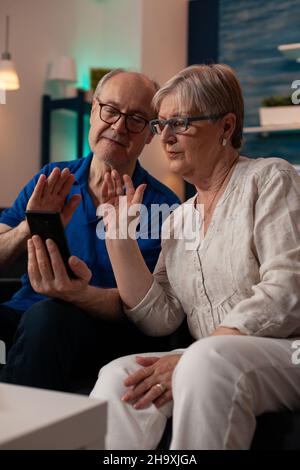 Pensionierter Mann und Frau plaudern auf einer Videokonferenz mit Freunden und Familie, während sie auf der Wohnzimmercouch sitzen. Gealtertes Paar, das zu Hause die Online-Fernkommunikation nutzt Stockfoto