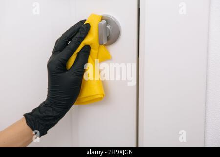 Reinigen des Türgriffs mit gelben Wischtüchern in schwarzen Handschuhen. Frau Hand mit Handtuch zum Reinigen. Desinfektion im Krankenhaus und öffentlichen Räumen gegen Corona Stockfoto