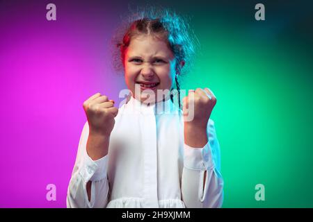 Portrait von wütenden kleinen Mädchen, Kid clenches Fäuste isoliert auf Farbverlauf lila blauen Hintergrund in Neonlicht Stockfoto