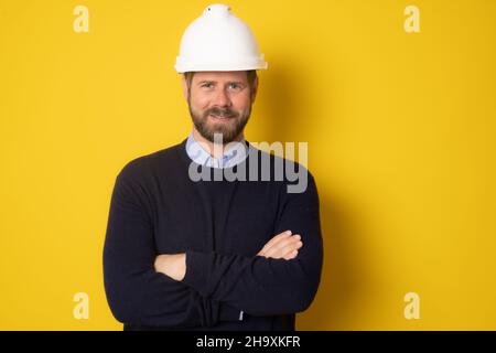 Junger Architekt Mann trägt Auftragnehmer Helm mit Armen über isolierten gelben Hintergrund gefaltet Stockfoto