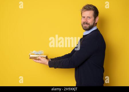 Junger, hübscher, blonder Mann mit Bart, der ein Geburtstagsgeschenk auf einem isolierten gelben Hintergrund hält Stockfoto